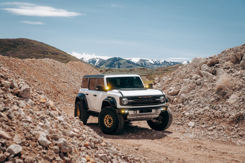 BRONCO RAPTOR driving on a rocky dirt road with mountains in the background with 38" Nitto Tires, Method gold wheels and Baja designs squadron lights